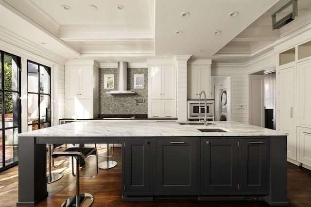 31 Lafayette Dr NE, Atlanta, GA 30309 - kitchen with stacked washing machine and dryer, wall chimney exhaust hood, white cabinetry, a large island with sink, and crown molding