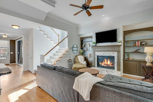 living room with crown molding, light wood-type flooring, built in features, ceiling fan, and a fireplace