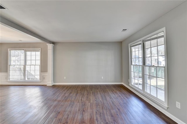 empty room featuring visible vents, baseboards, wainscoting, dark wood finished floors, and decorative columns