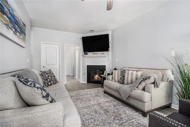 living area with a textured ceiling, ceiling fan, a glass covered fireplace, and wood finished floors