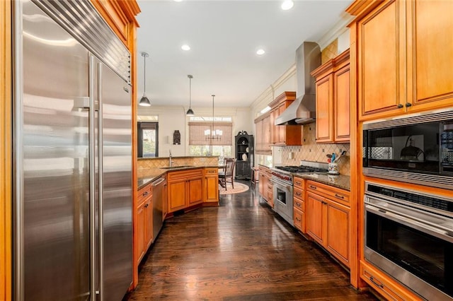 kitchen featuring wall chimney exhaust hood, decorative light fixtures, built in appliances, stone counters, and a notable chandelier