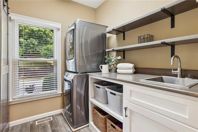 laundry room with a sink, stacked washing maching and dryer, baseboards, wood finished floors, and visible vents