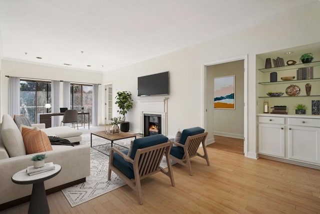 living area featuring built in shelves, baseboards, light wood-style floors, a lit fireplace, and ornamental molding