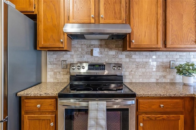 kitchen with tasteful backsplash, appliances with stainless steel finishes, brown cabinets, light stone counters, and under cabinet range hood