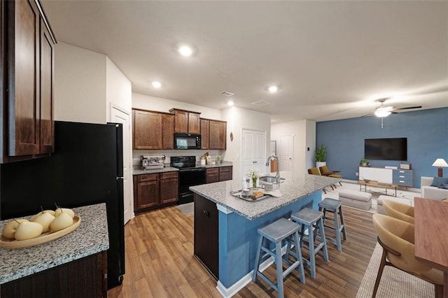 kitchen with ceiling fan, a kitchen island with sink, black appliances, light hardwood / wood-style flooring, and a breakfast bar area