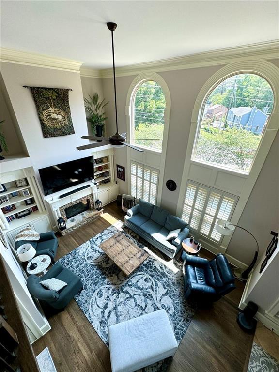 living room with a stone fireplace, a high ceiling, a ceiling fan, ornamental molding, and dark wood finished floors