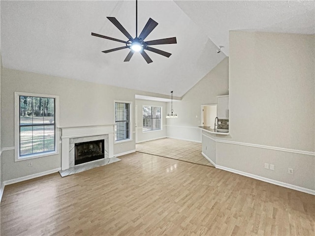 unfurnished living room with high vaulted ceiling, light wood-type flooring, ceiling fan with notable chandelier, and a high end fireplace