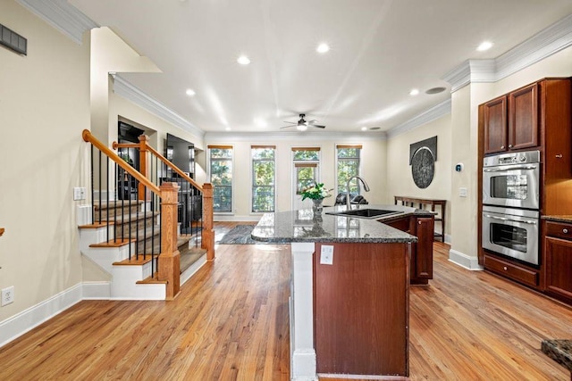 kitchen with ornamental molding, stainless steel double oven, a kitchen island with sink, sink, and light hardwood / wood-style floors