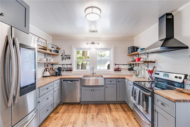 kitchen with stainless steel appliances, wall chimney exhaust hood, wooden counters, sink, and light hardwood / wood-style flooring