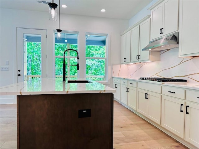 kitchen with light wood-style flooring, under cabinet range hood, a sink, tasteful backsplash, and gas stovetop