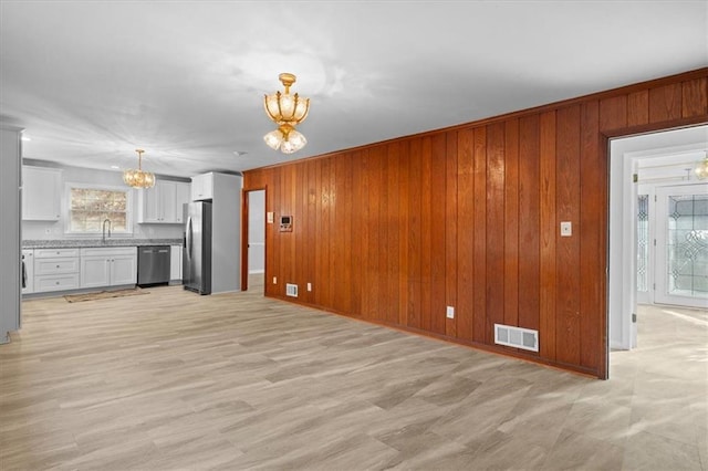 unfurnished living room with light wood finished floors, wooden walls, visible vents, a sink, and a notable chandelier