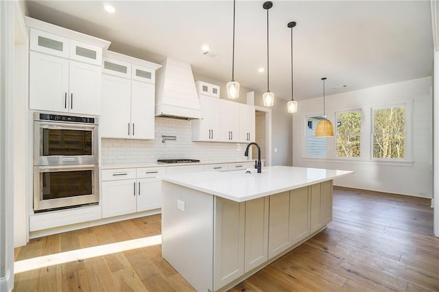 kitchen with a sink, double oven, custom exhaust hood, gas cooktop, and a kitchen island with sink