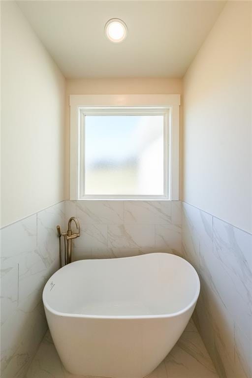 bathroom with marble finish floor, wainscoting, a soaking tub, and tile walls