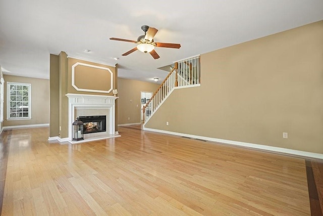 unfurnished living room with baseboards, a ceiling fan, a glass covered fireplace, stairs, and light wood-style floors