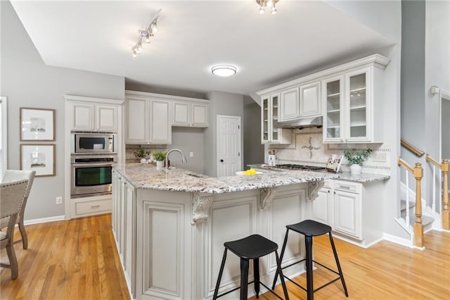 kitchen with a sink, under cabinet range hood, backsplash, glass insert cabinets, and appliances with stainless steel finishes