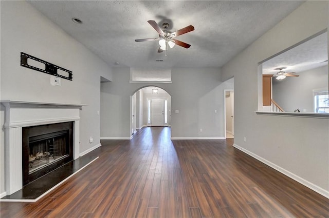 unfurnished living room with baseboards, arched walkways, a fireplace with raised hearth, a ceiling fan, and dark wood-style floors