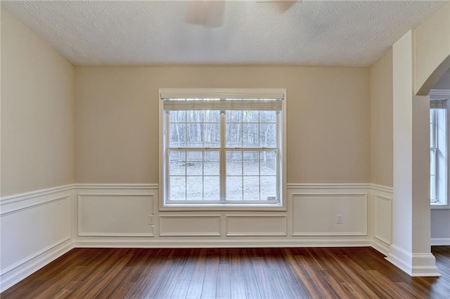 empty room featuring arched walkways, dark wood-type flooring, a textured ceiling, and a wainscoted wall