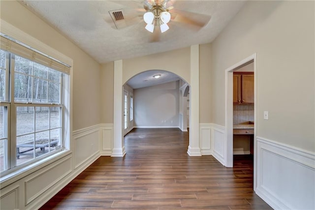 unfurnished dining area with arched walkways, a ceiling fan, wainscoting, dark wood-style floors, and a decorative wall