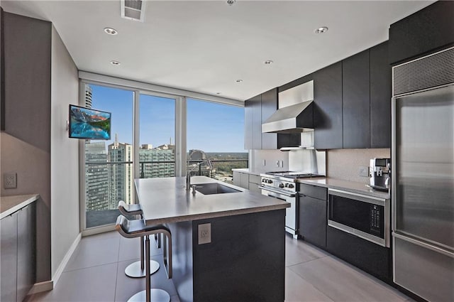 kitchen featuring wall chimney range hood, sink, built in appliances, an island with sink, and a wealth of natural light