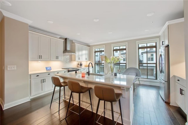 kitchen featuring white cabinetry, an island with sink, wall chimney exhaust hood, ornamental molding, and stainless steel fridge