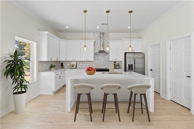 kitchen with stainless steel refrigerator with ice dispenser, white cabinets, a kitchen island with sink, and wall chimney exhaust hood