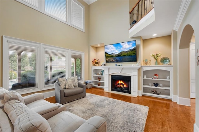 living area featuring arched walkways, a fireplace with flush hearth, wood finished floors, crown molding, and built in shelves