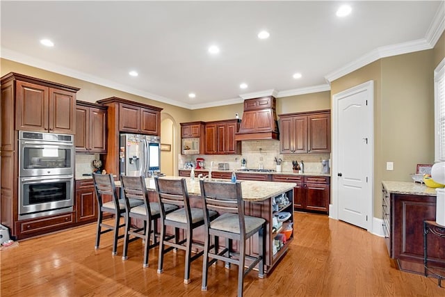 kitchen with arched walkways, appliances with stainless steel finishes, a breakfast bar, a kitchen island with sink, and custom exhaust hood