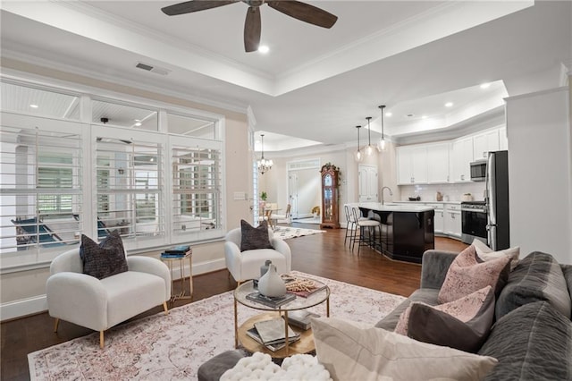 living room featuring sink, dark wood-type flooring, a tray ceiling, ceiling fan with notable chandelier, and ornamental molding