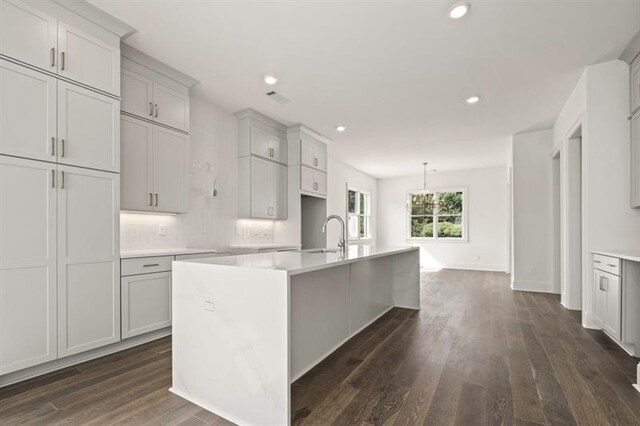 kitchen with tasteful backsplash, hanging light fixtures, sink, a center island with sink, and dark wood-type flooring