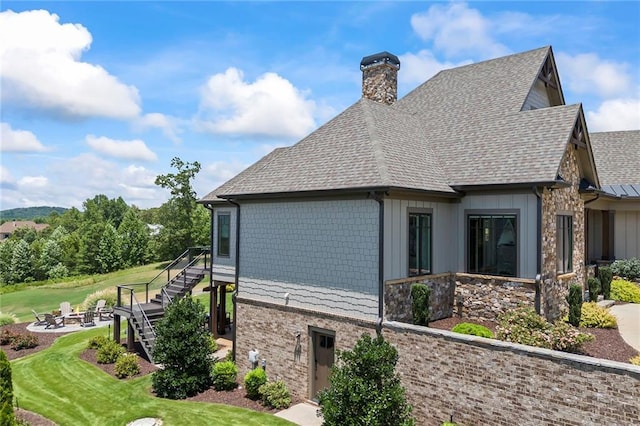 view of side of property featuring a chimney, a shingled roof, a lawn, an outdoor fire pit, and stairway
