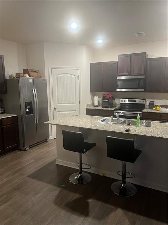 kitchen featuring a breakfast bar area, appliances with stainless steel finishes, dark brown cabinets, dark hardwood / wood-style floors, and light stone counters