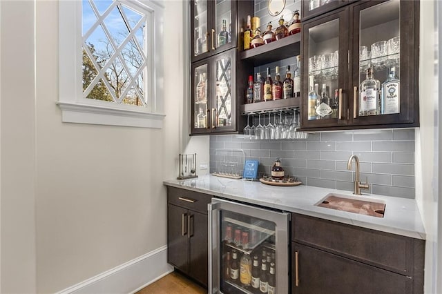 bar with sink, wine cooler, tasteful backsplash, dark brown cabinets, and light stone counters