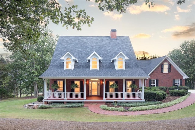 new england style home featuring a porch, a yard, brick siding, and a shingled roof