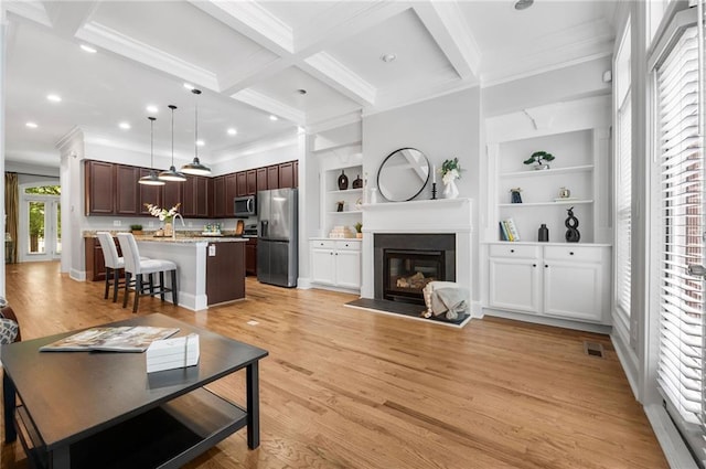 living area with ornamental molding, beam ceiling, a glass covered fireplace, light wood-style floors, and built in shelves