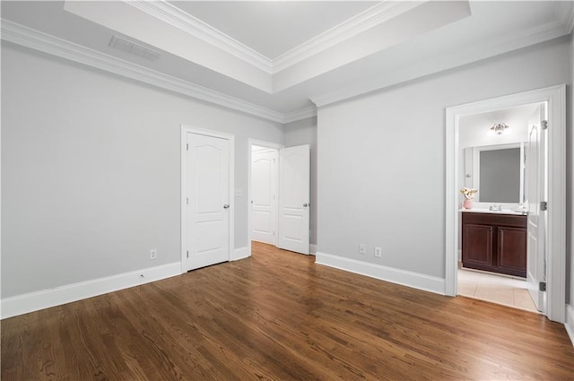 unfurnished bedroom featuring wood finished floors, a tray ceiling, crown molding, baseboards, and ensuite bath