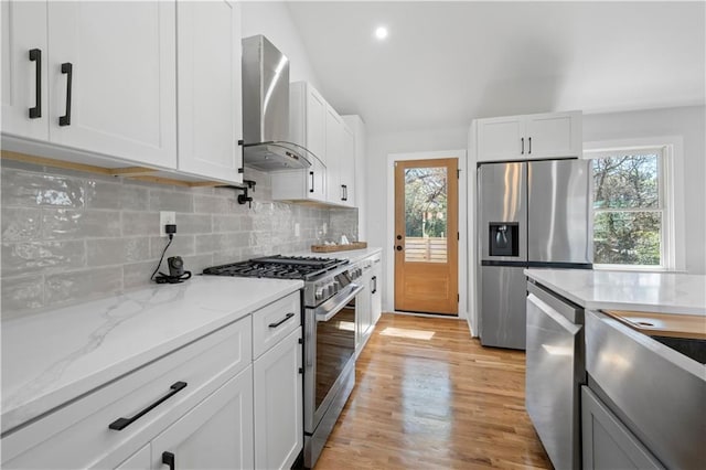 kitchen with decorative backsplash, appliances with stainless steel finishes, light wood-type flooring, wall chimney range hood, and white cabinetry