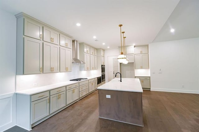 kitchen featuring wall chimney exhaust hood, gas stovetop, a kitchen island with sink, dark wood-type flooring, and decorative light fixtures