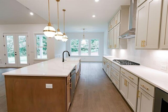 kitchen featuring plenty of natural light, wall chimney range hood, an island with sink, decorative light fixtures, and stainless steel appliances