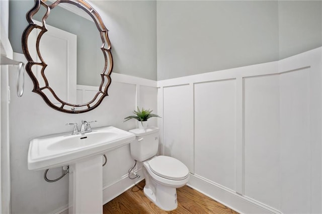 half bathroom featuring toilet, a decorative wall, wood finished floors, and wainscoting