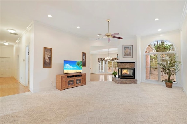 living room featuring light carpet, a stone fireplace, crown molding, and ceiling fan with notable chandelier