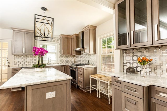 kitchen with glass insert cabinets, a center island, decorative light fixtures, stainless steel stove, and wall chimney range hood