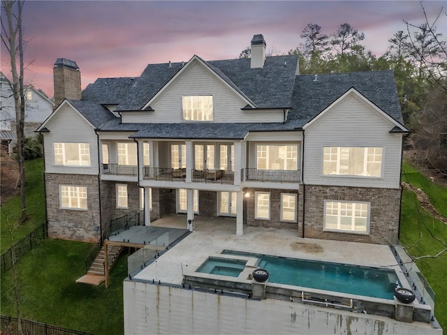 back of house at dusk with a fenced backyard, a lawn, a balcony, a pool with connected hot tub, and a patio