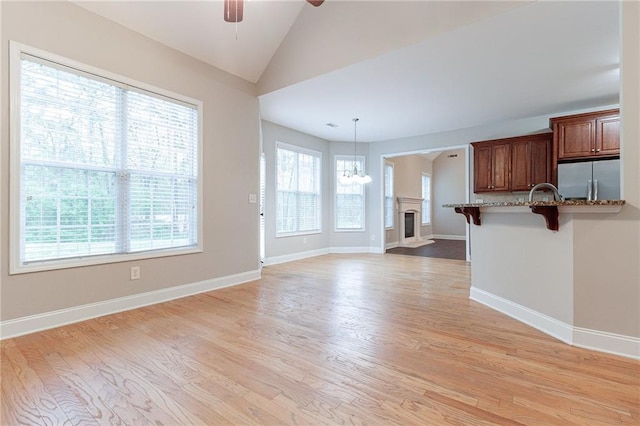 unfurnished living room with ceiling fan with notable chandelier, vaulted ceiling, a wealth of natural light, and light hardwood / wood-style flooring