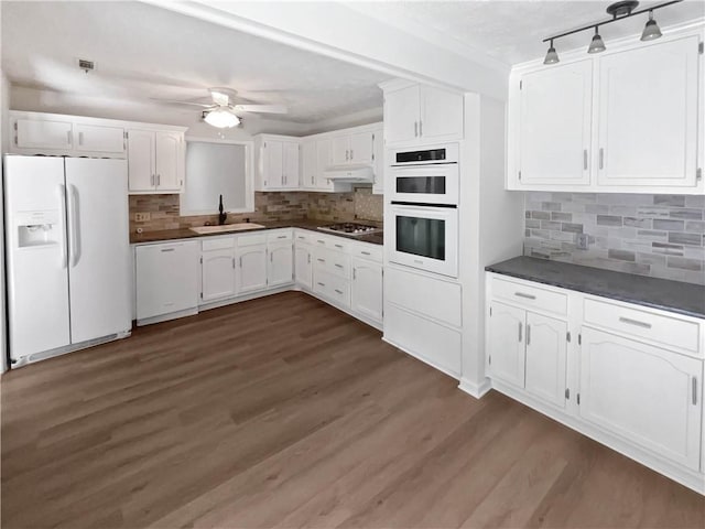kitchen featuring white appliances, white cabinetry, dark countertops, under cabinet range hood, and a sink