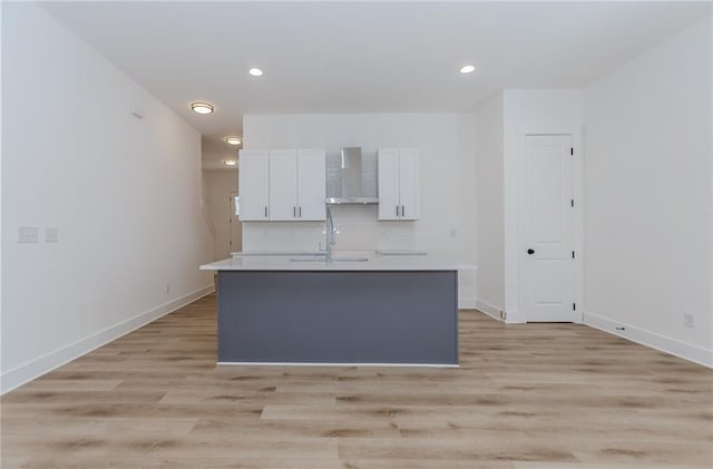 kitchen with a kitchen island with sink, a sink, white cabinetry, backsplash, and wall chimney exhaust hood