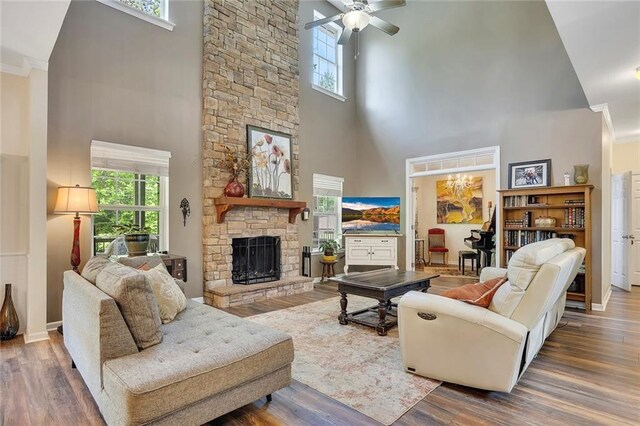living area with a towering ceiling, a stone fireplace, healthy amount of natural light, a ceiling fan, and wood finished floors