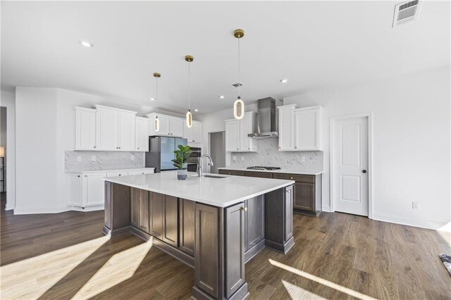 kitchen featuring wall chimney exhaust hood, hanging light fixtures, a large island with sink, stainless steel appliances, and white cabinets