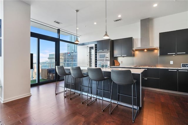 kitchen featuring visible vents, wall chimney exhaust hood, stainless steel microwave, dark cabinets, and a wall of windows