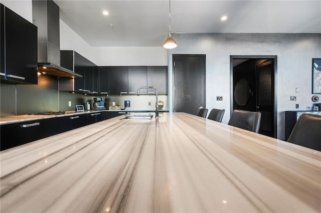 kitchen featuring wall chimney exhaust hood, dark cabinets, light countertops, pendant lighting, and a sink