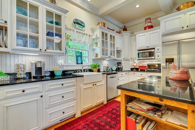 kitchen with built in appliances, beamed ceiling, dark stone counters, sink, and white cabinetry